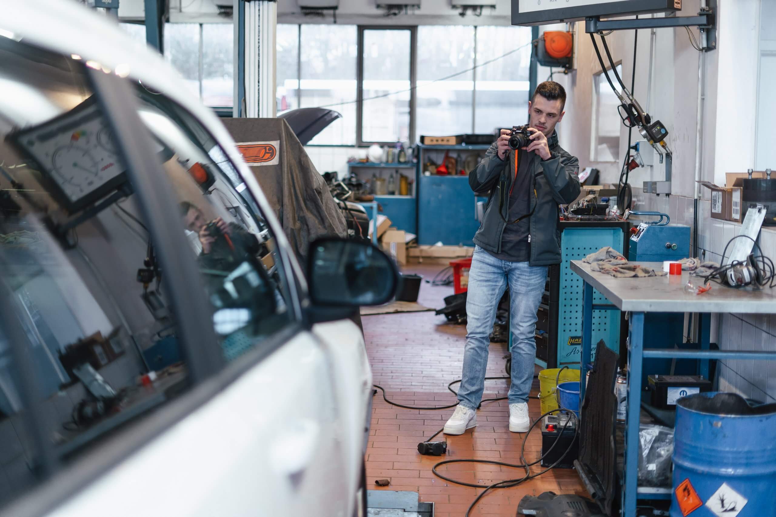 Ein Mann fotografiert in einer Werkstatt ein Auto mit einer Kamera, umgeben von Werkzeugen und Arbeitsgeräten