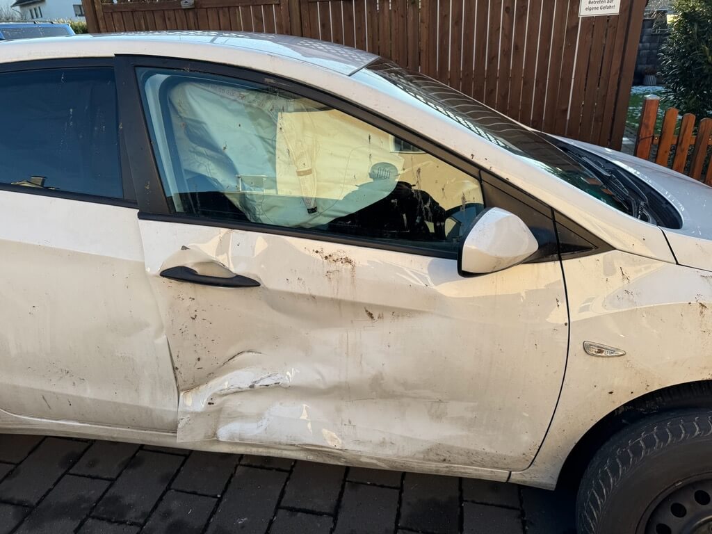 Ein weißes Auto mit sichtbaren Schäden an den Seiten, Schmutzspuren und einem ausgelösten Airbag steht auf dem Asphalt neben einem Holzzaun
